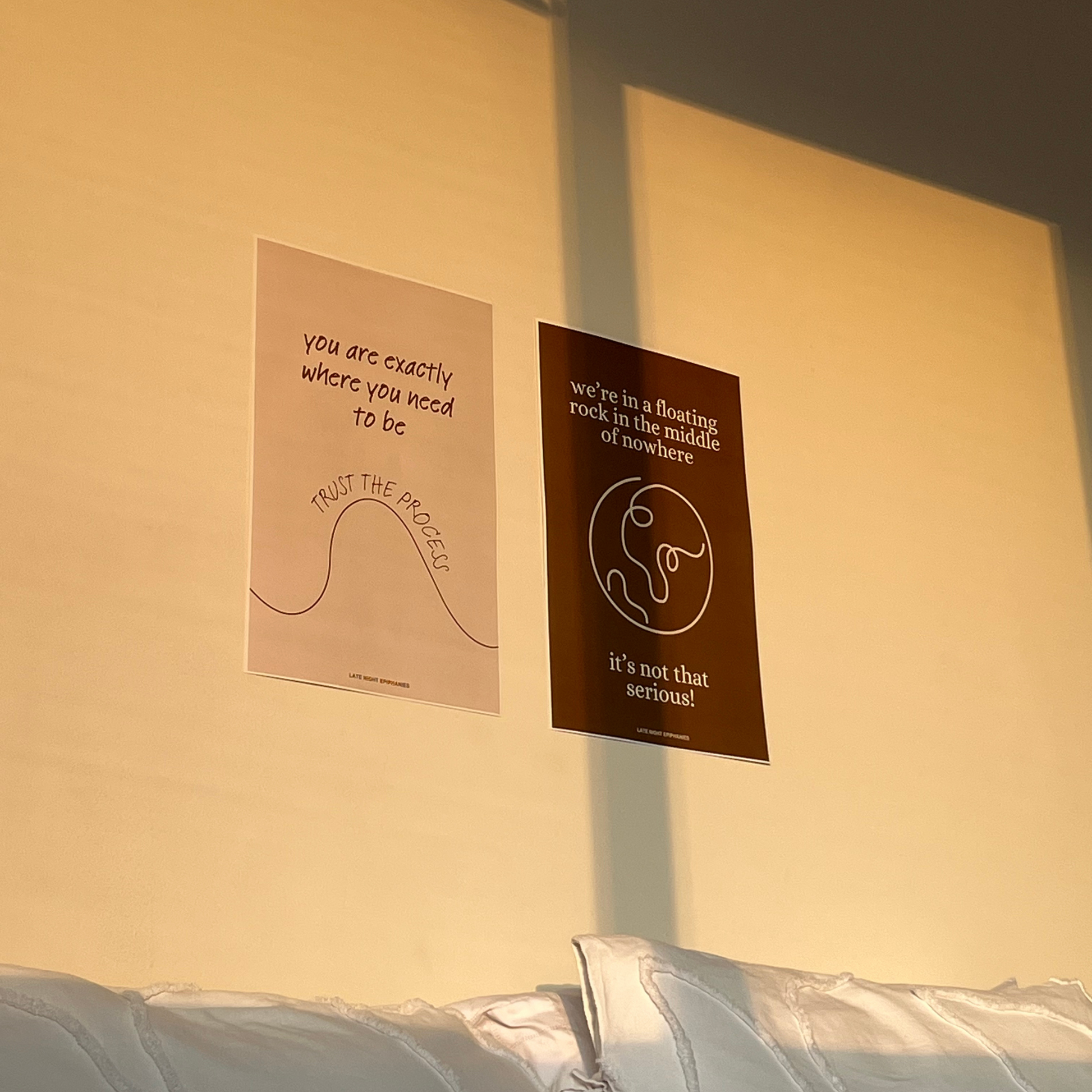 Image of two posters in a bedroom wall during golden hour. One is beige and says "you are exactly where you need to be, trust the process." The other is brown and says "we're in a floating rock in the middle of nowhere. it's not that serious!"